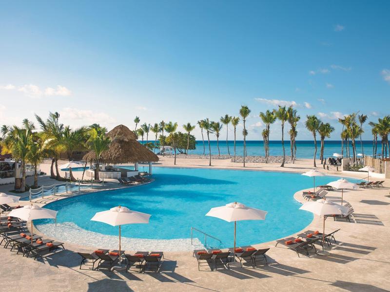 Large round pool at sunny resort with loungers, umbrellas, and palm trees on the beach.