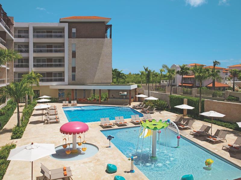 Modern outdoor pool with kids' play area, surrounded by loungers and palm trees under a blue sky.
