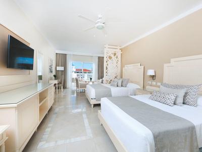 Helles Hotelzimmer mit zwei Doppelbetten, Fernseher, Ventilator und Balkon mit Meeresblick.