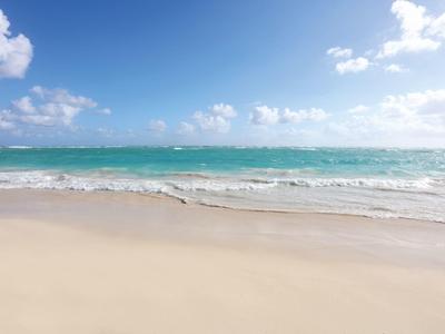 Weißer Sandstrand mit sanften Wellen und klarem blauem Himmel mit wenigen Wolken.