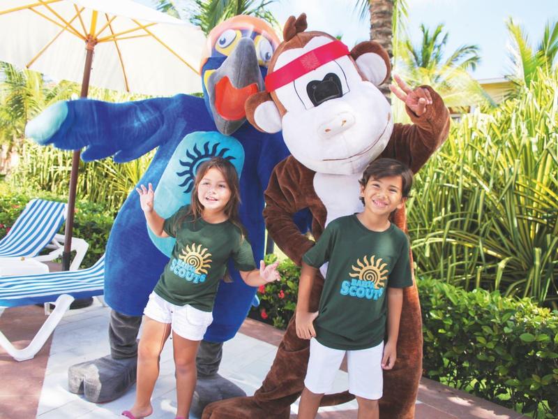 Zwei Kinder in grünen Shirts posieren mit einem blauen Papagei- und einem braunen Affenkostüm im Freien.