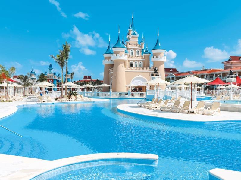 Großer Pool mit klarem Wasser, Sonnenschirmen und Schloss im Hintergrund unter blauem Himmel.
