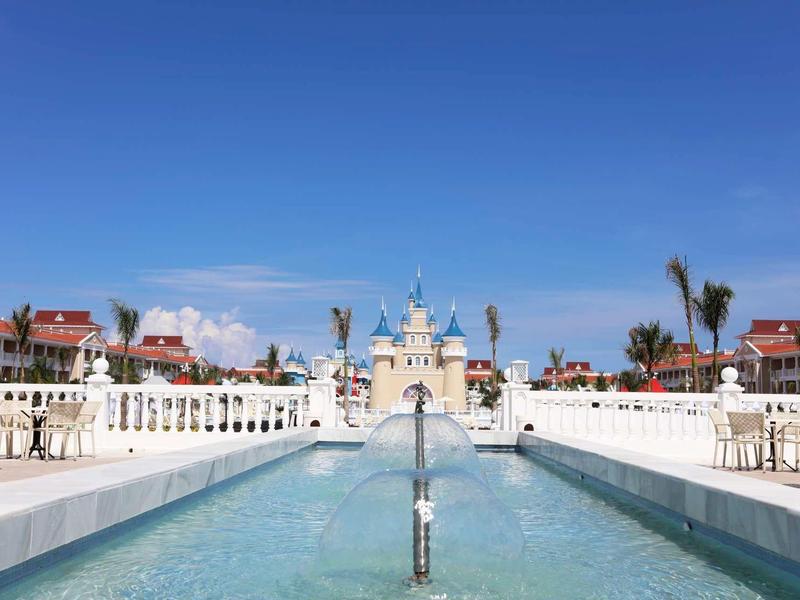 Großes Schloss mit Wasserfontäne und blauen Himmel, flankiert von weißen Zäunen und Gebäuden.