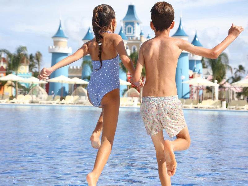 Zwei Kinder laufen am Poolufer, hinter ihnen ein blauer Wasserpark mit Türmen.