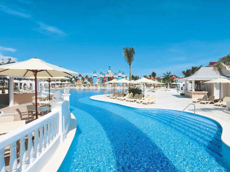 Großer blauer Pool mit weißem Rand, Sonnenschirmen und Palmen unter strahlend blauem Himmel.