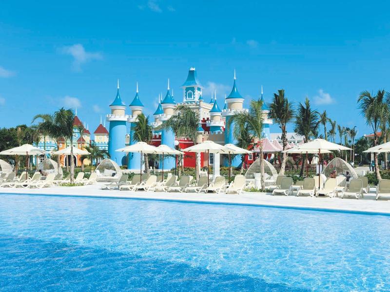 Großes Schwimmbecken mit blauem Wasser vor einem märchenhaften blauen Schloss und Sonnenschirmen.