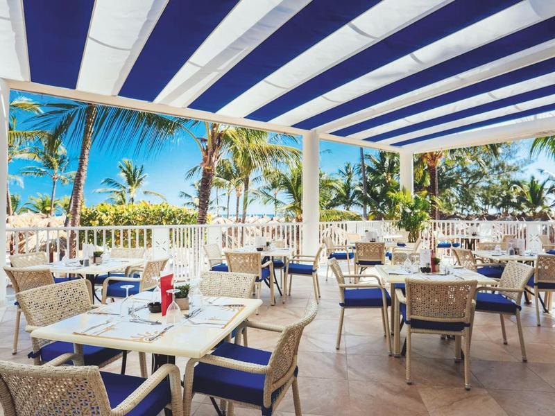 Freiluftrestaurant mit blauen Sonnensegeln, Flechtstühlen und tropischer Palmenkulisse.