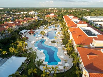 Blick auf einen Hotelkomplex mit großem Pool, umgeben von Sonnenschirmen und Palmen.
