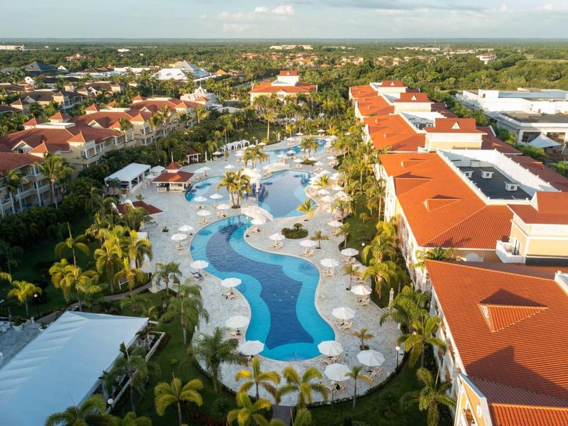 Widok z lotu ptaka na hotel z dużym basenem i leżakami, otoczony palmami i budynkami z czerwonymi dachami.