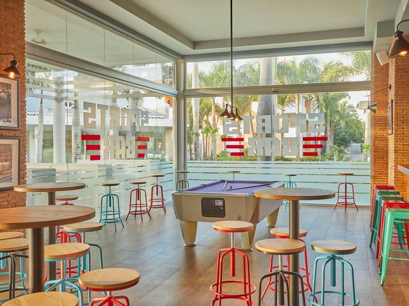 Jasno urządzone miejsce rekreacyjne z bilardem i stołkami barowymi przy oknie z widokiem na palmy.