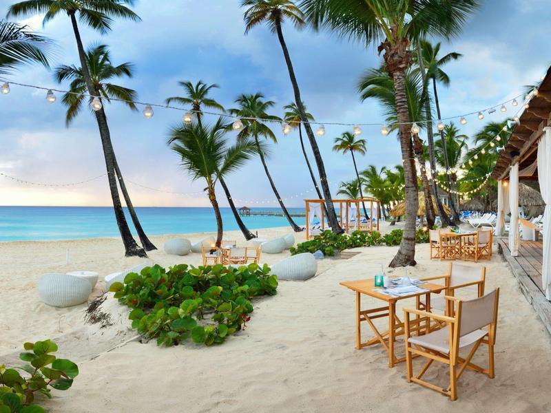 Beach with palm trees, sand, and tables at an open restaurant with sea view.