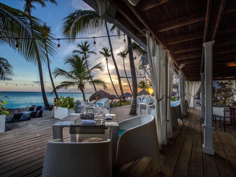 Beachside restaurant at sunset with tables, chairs, and palm trees by the sea.