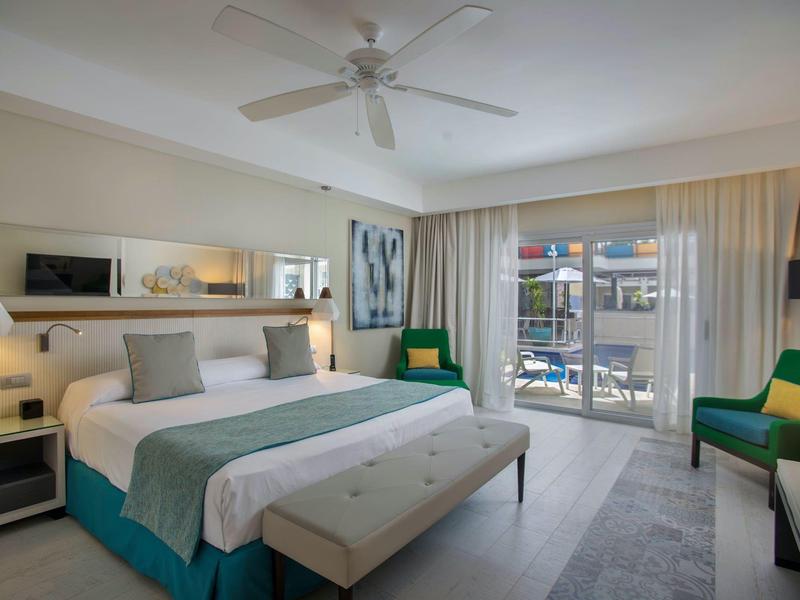 Modern hotel room with large bed, seating area, and balcony overlooking outdoors.