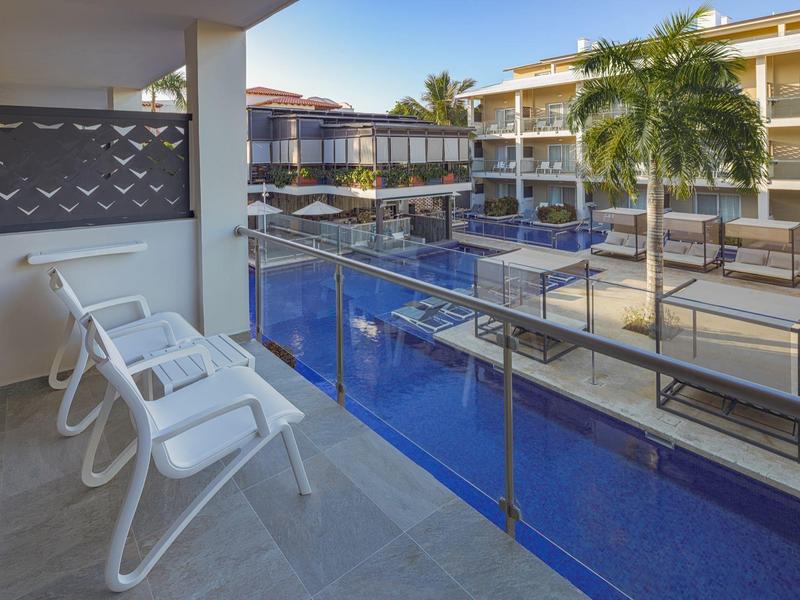 Balcony with white chairs overlooking a hotel pool with palm trees and multiple buildings.