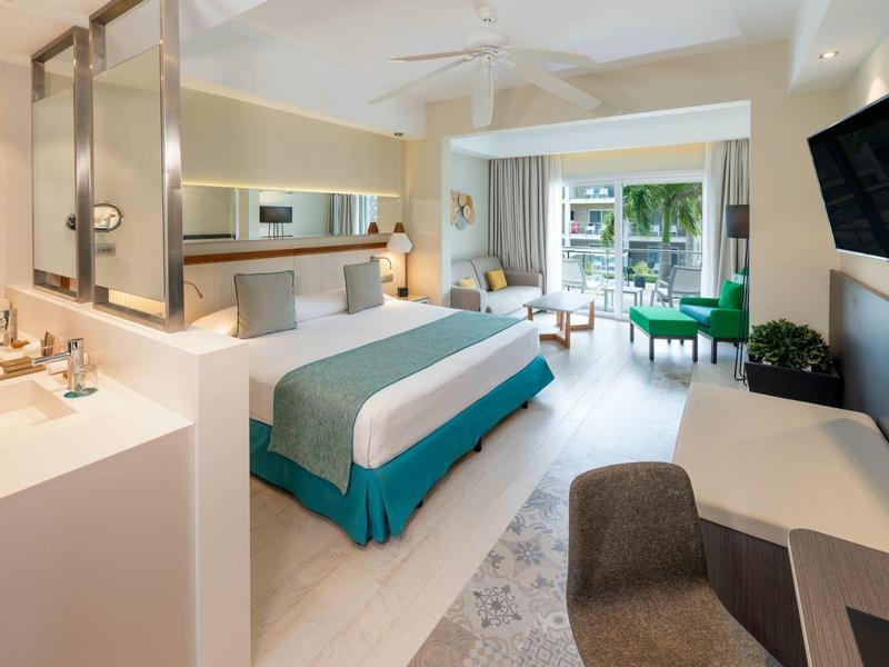 Bright hotel room with bed, desk, sofa, and balcony overlooking garden