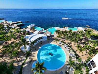 Blick auf großen Pool mit Palmen, weißen Gebäuden und blauem Meer im Hintergrund, sonniger Tag.