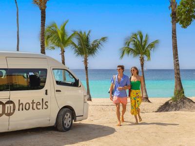 Rodina kráčí po písečné pláži kolem bílého dodávky s logem hotelu, palmy a moře v pozadí.