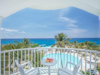 Balkon s bílým zábradlím, stůl s ovocem, výhled na bazén a moře pod modrou oblohou.