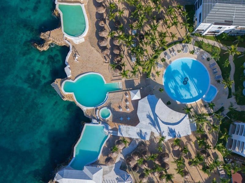 Letecký pohled na resort s několika bazény u moře a palmami.