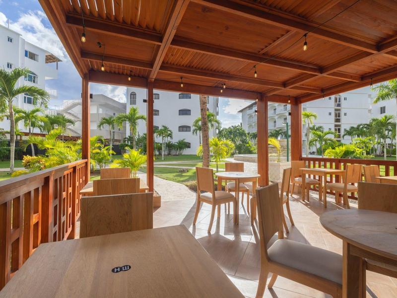 Zastřešená terasa s dřevěnými stoly a židlemi, obklopená zelení a hotelovými budovami.