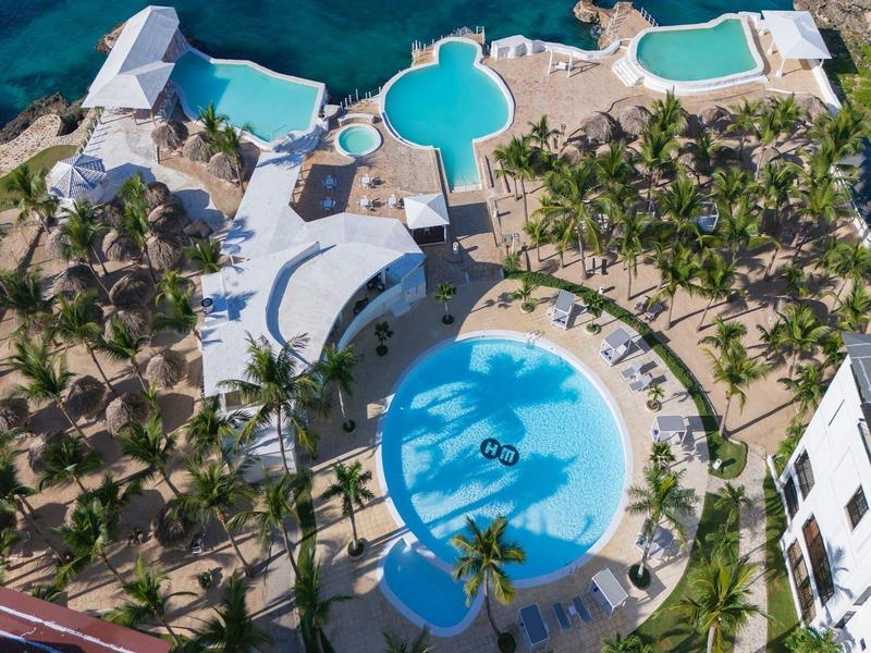 Letecký pohled na hotelový bazén s palmami a lehátky v tropickém prostředí.