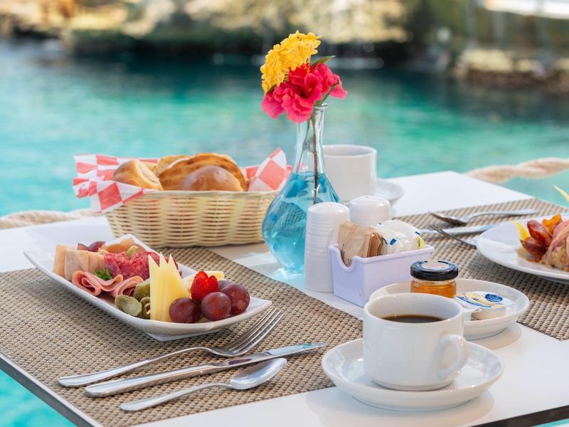 Stůl na snídani s kávou, pečivem a ovocem u vody s výhledem na pobřeží.
