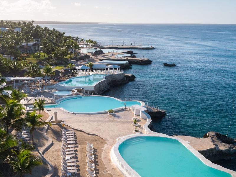 Pohled na resort s několika bazény u pobřeží a lehátky obklopený palmami.