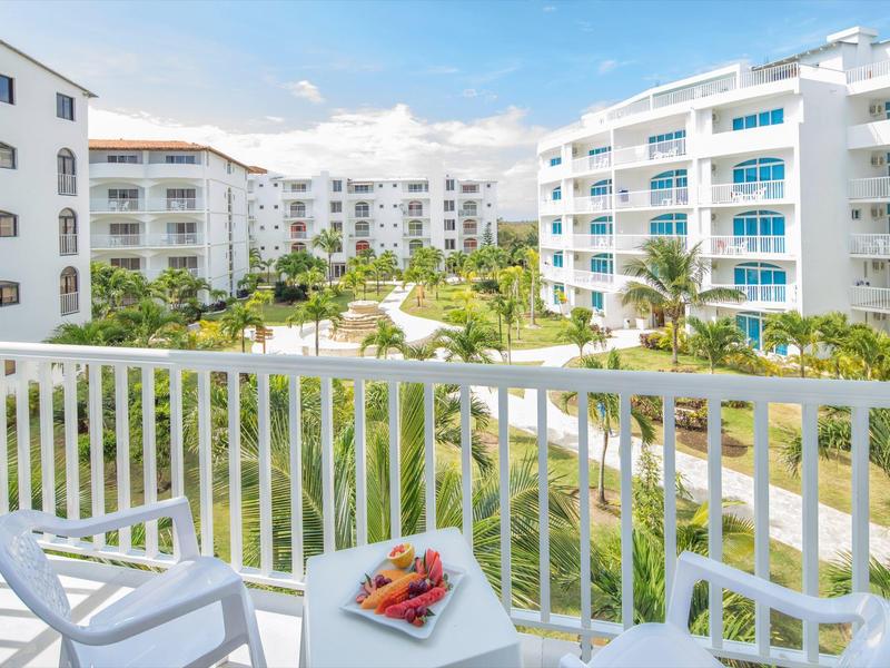 Výhled z balkonu na moderní hotely a tropickou zahradu za slunečného dne.