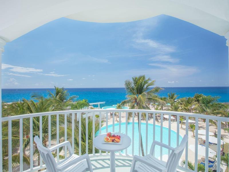 Balkon s bílým zábradlím, stůl s ovocem, výhled na bazén a moře pod modrou oblohou.