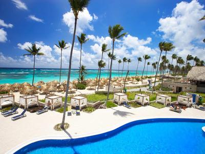 Plážové letovisko s bazénem, lehátky a palmami u tyrkysového moře pod modrou oblohou.