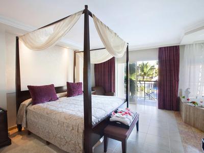 Elegancki pokój hotelowy z łóżkiem z baldachimem, fioletowymi zasłonami i balkonem z widokiem na morze.