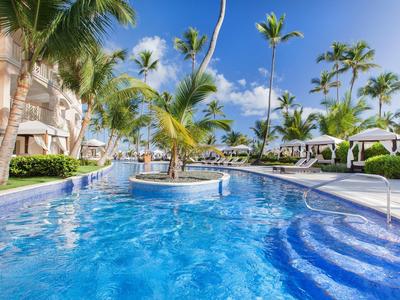 Großer Pool mit blauer Wasseroberfläche, Palmen und Liegen unter blauem Himmel in Tropenanlage.