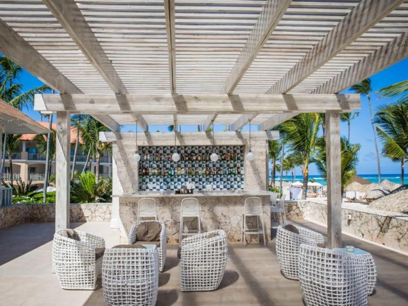 Otwarta przestrzeń wypoczynkowa z meblami z rattanu pod pergolą przy plaży z roślinami i widokiem na morze.