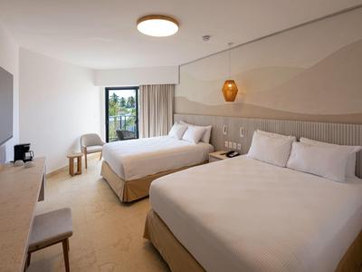 Camera d'hotel luminosa con due letti, scrivania, sedie e balcone con vista sul verde.
