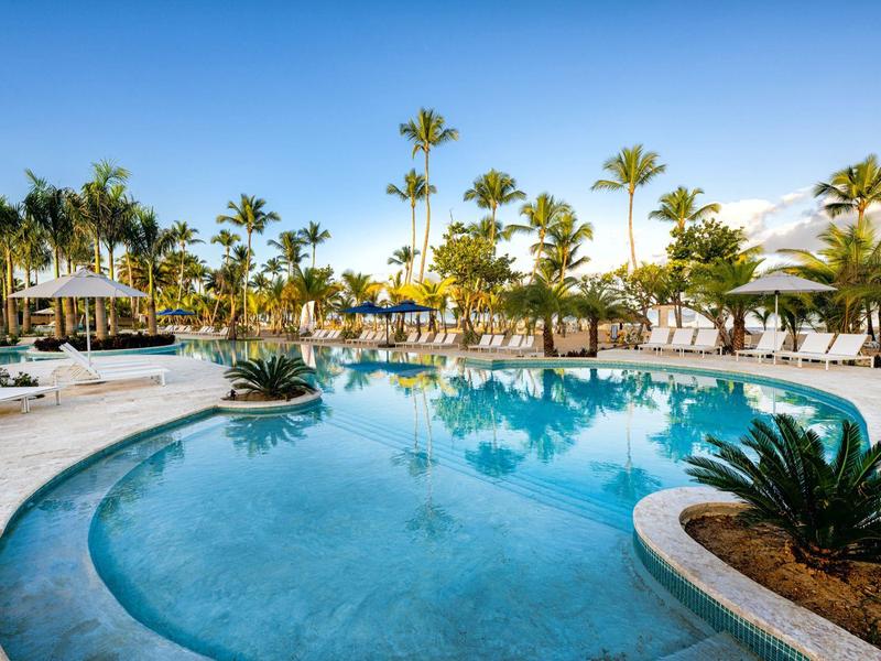 Grande piscina rotonda circondata da palme e lettini sotto un cielo sereno.