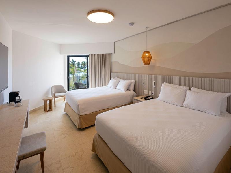 Camera d'hotel luminosa con due letti, scrivania, sedie e balcone con vista sul verde.