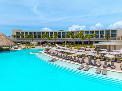 Modern hotel complex with large pool, sun loungers, and umbrellas under a blue sky.