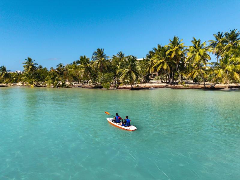 Zwei Personen paddeln auf einem Kajak in türkisfarbenem Wasser vor Palmenstrand.