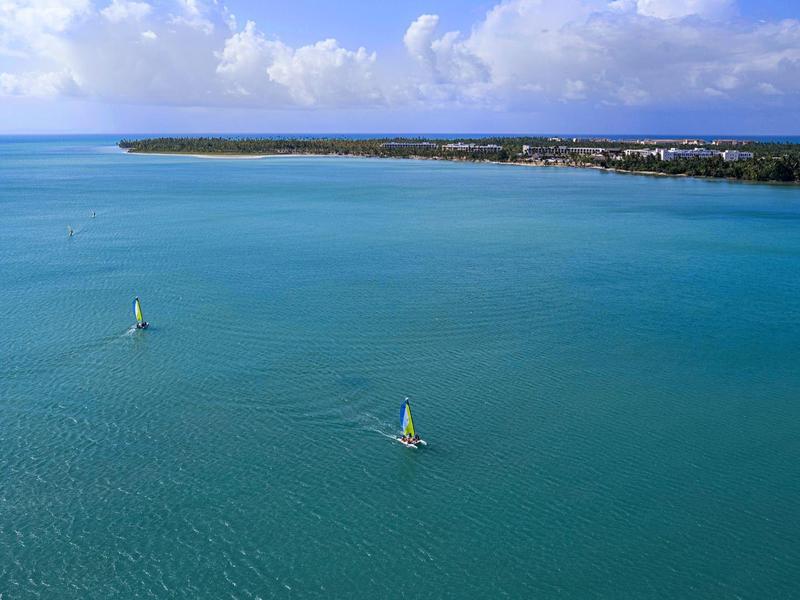 Blick auf klares blaues Wasser mit zwei Segelbooten und einer Baumküste unter bewölktem Himmel.