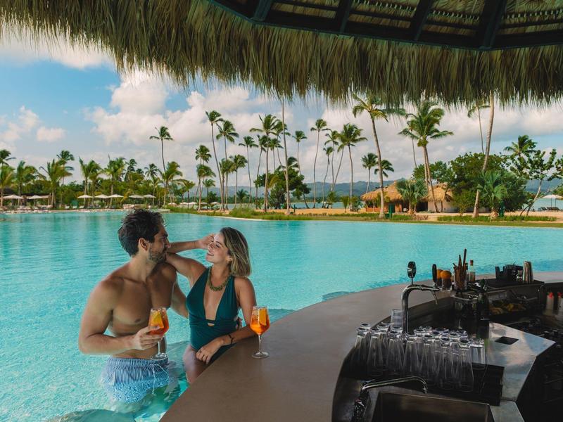 Paar sitzt an einer schwimmenden Bar im Pool mit Palmen und blauem Himmel im Hintergrund.