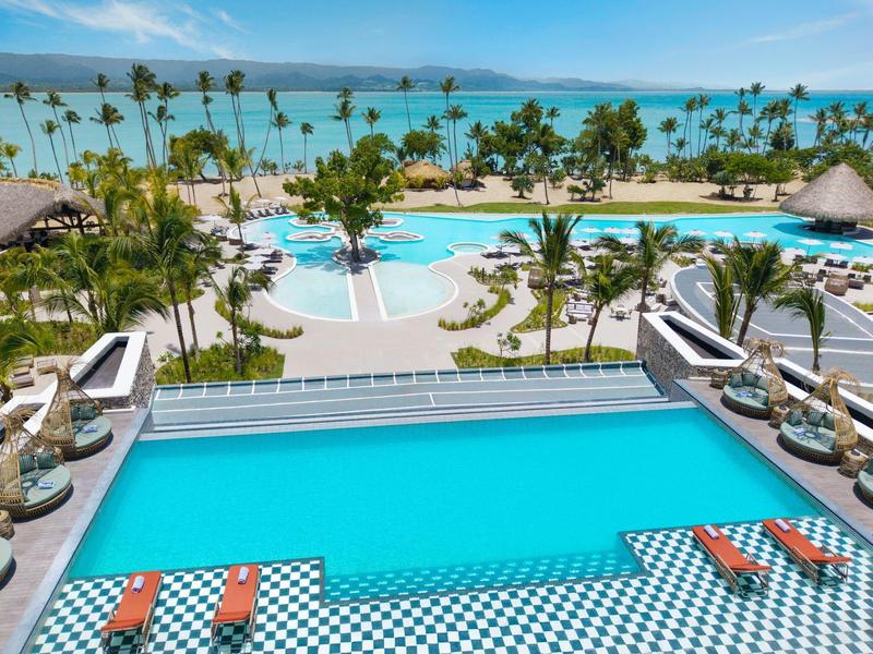 Blick auf einen Poolbereich mit Liegen und Palmen neben dem Meer an einem sonnigen Tag.