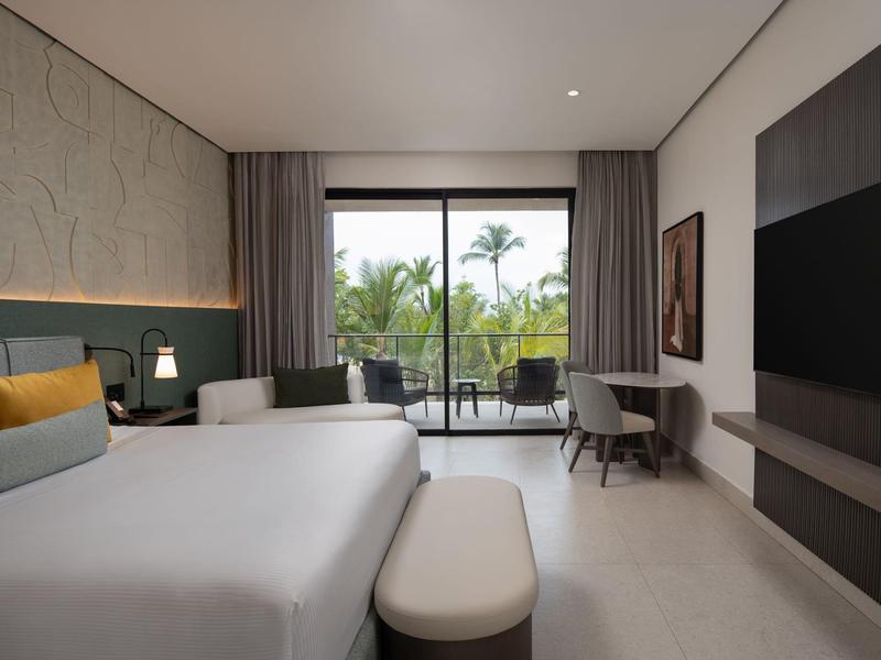 Modernes Hotelzimmer mit Doppelbett, Fensterfront und Balkon mit Blick auf Palmen.