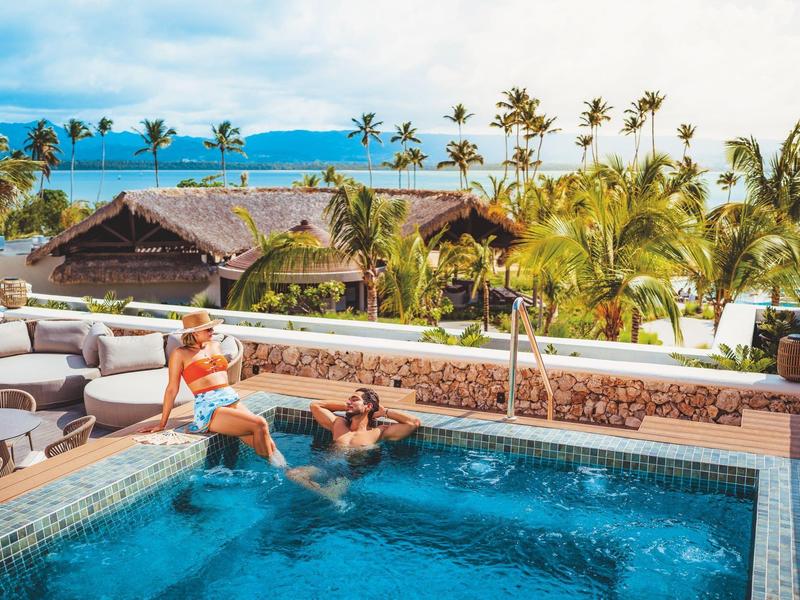 Zwei Personen entspannen im Pool mit Blick auf tropische Palmen und das Meer.