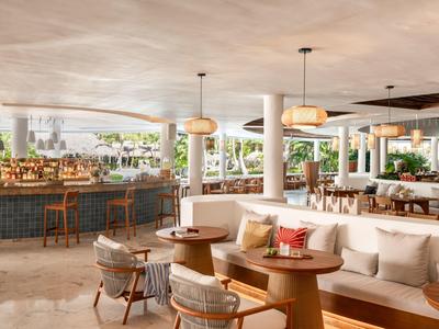 Modern open-air hotel lounge with wooden tables, cushioned seating, and bar stools.