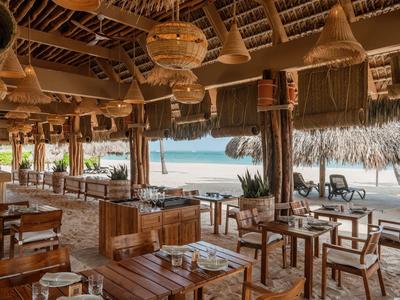 Beachside restaurant with wooden tables, chairs, woven lamps, and ocean views under a thatched roof.