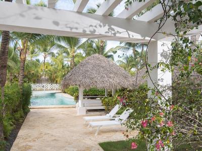 Poolside lounge chairs under a thatched umbrella near a tropical pool surrounded by palm trees.