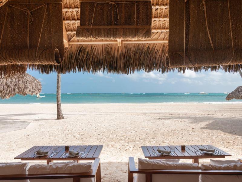Thatched roof shades two wooden tables set for dining on a pristine white sandy beach by the turquoise sea.