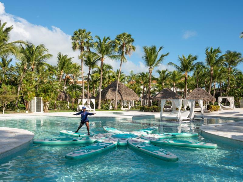 Resort pool with palm trees, kayaks, and a person balancing on a floating platform under blue sky.
