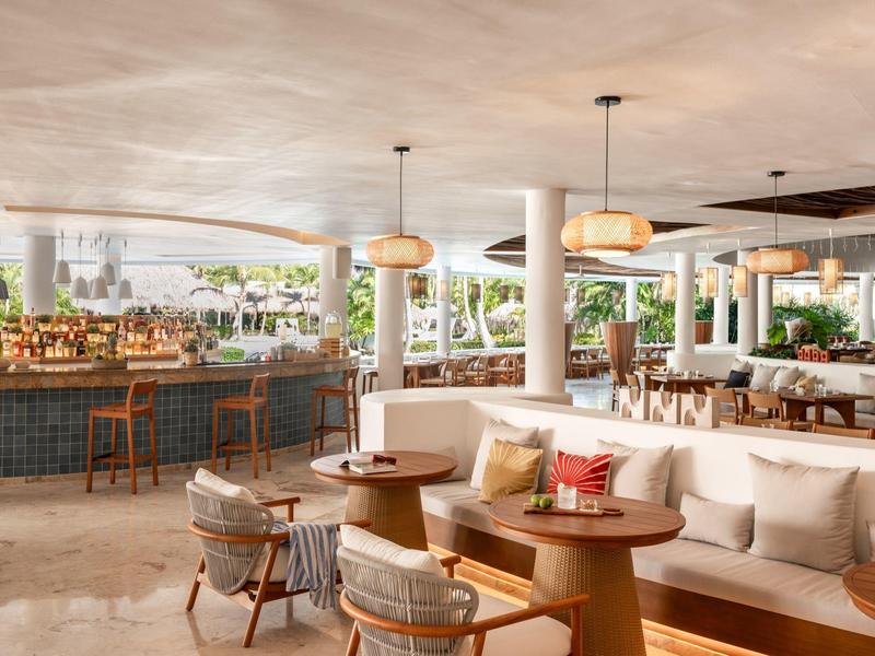 Modern open-air hotel lounge with wooden tables, cushioned seating, and bar stools.