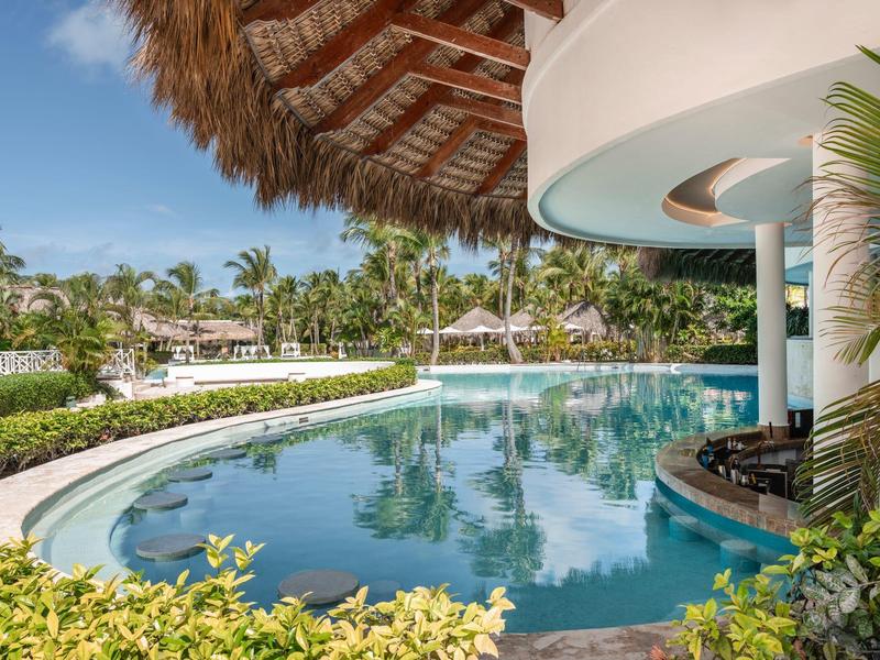 Curved pool under thatched roof with tropical gardens and palm trees in the background.